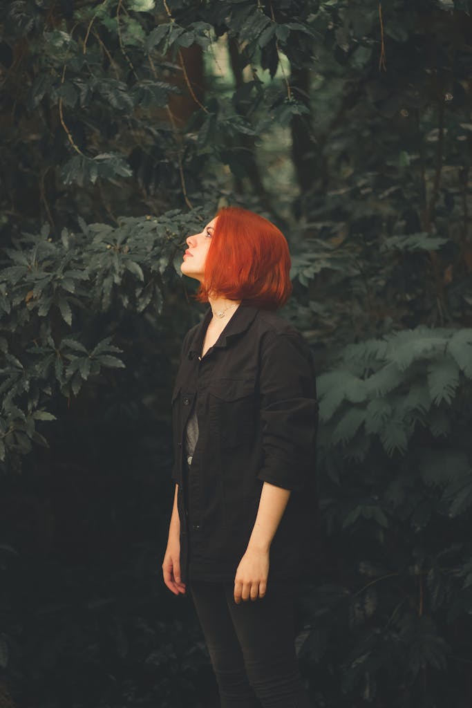 Woman standing amidst dark green plants, looking up thoughtfully in an outdoor setting.